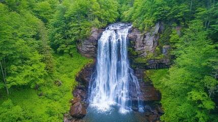 Aerial view of majestic waterfall cascading down a rocky cliff, surrounded by vibrant forest greenery.