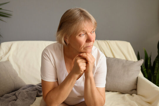Thoughtful senior woman sitting alone on sofa looking pensive and worried elderly retired female with sad expression in her home leaning on her hands looking away
