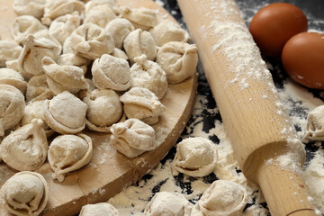 Raw dumplings, selective focus. Cooking pelmeni, flour, eggs and wooden rolling pin. Meat pelmeni or ravioli on wooden board, dark table. Slavic traditional dish. Food background