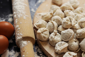 Raw dumplings, selective focus. Cooking pelmeni, flour, eggs and wooden rolling pin. Meat pelmeni or ravioli on wooden board, dark table. Slavic traditional dish. Food background
