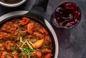 Sizzling dish of mixed beef meats, spiced sauce, garnished with fresh herbs. In the background, a textured glass filled with a deep red drink, alongside a copper bowl of fresh ginger.