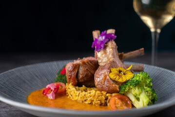 Beautifully plated dish of lamb chops garnished with edible flowers, accompanied by colorful vegetables and a rich sauce, a glass of white wine to the side. © alimrannahid