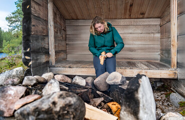 Outdoor woman sitting in a rustic log shelter hut (slogbod) in the Swedish nature, skillfully...