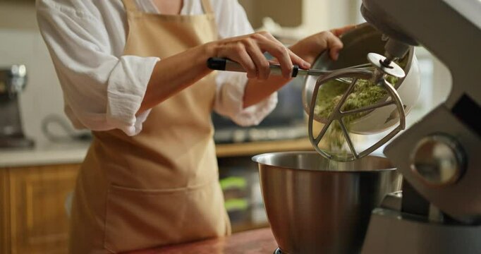 Confident girl Baker in a beige apron pours green shortbread dough into a mixer while preparing desserts in a bakery