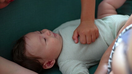 Baby lying on the couch, gazing up with a curious expression, while a child’s hand rests gently...