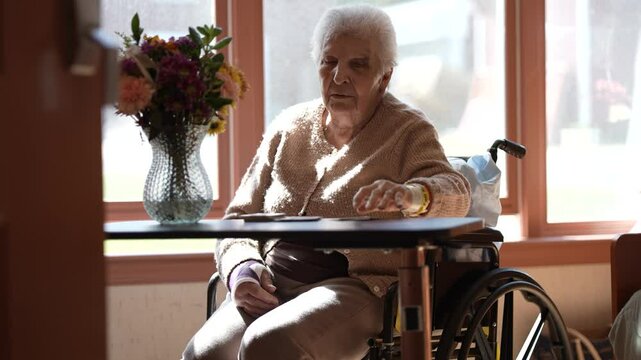 An elderly woman in a wheelchair is playing cards at a table. She is wearing a beige sweater and has her hand on a deck of cards. A vase with flowers is on the table next to her.