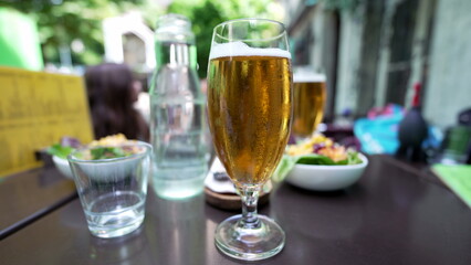 Close-up of a glass of beer with a blurred salad and a glass of water in the background on an outdoor restaurant table. A refreshing drink capturing the essence of a relaxing dining experience