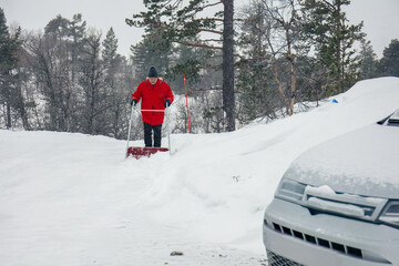 Man in red jacket pushing a snow pusher on a snow-covered road by mountain cabin ,winter task,...