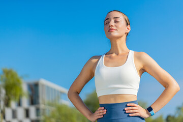 Fototapeta premium Young Caucasia female runner is short of breath after a fast sprint run in urban city park. Fit female athlete with hands on hips looking around and breathes deeply on summer day. Healthy lifestyle.
