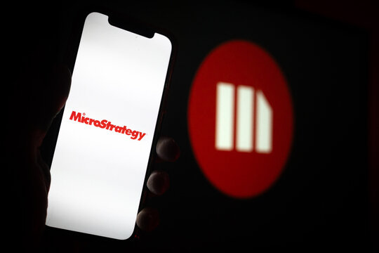 Silhouette of Hand Holding iPhone with MicroStrategy Logo Icon in Front of Computer Screen Displaying Logo &ndash; Business Intelligence and Analytics Solutions &ndash; MicroStrategy Incorporated (MSTR)
