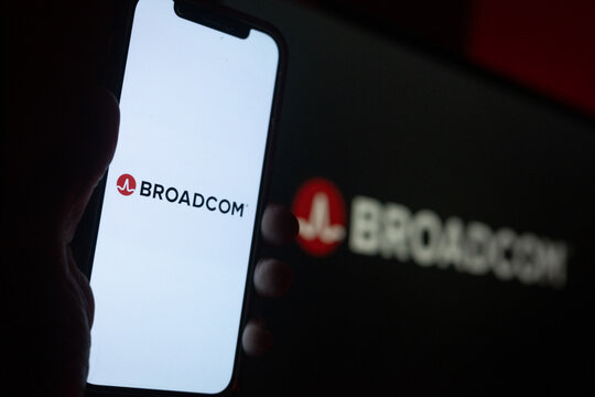 Silhouette of Hand Holding iPhone with Broadcom Logo in Front of Computer Screen Displaying Logo &ndash; Semiconductor and Infrastructure Solutions &ndash; Broadcom Inc. (AVGO)