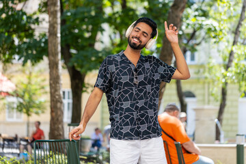 Happy relaxed young Indian man in wireless headphones listening favorite energetic disco music dancing outdoors. Handsome overjoyed tourist male Hispanic guy standing in city street park. Lifestyles.