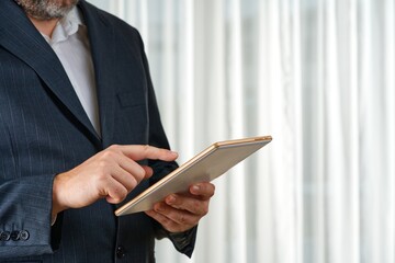 A confident businessman in a tailored suit stands in a modern office, intently using a tablet....
