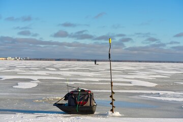 An ice fishing hole is drilled in the ice with a fishing rod in it. A sled with a tackle box and gear is nearby, while a person is seen ice fishing in the distance.