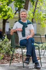 Fototapeta premium Portrait of happy bearded middle-aged smiling man glad expression looking at camera resting relaxation feel satisfied good news outdoors. Guy sitting on chair on urban city sunshine street park