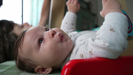 Close-up of baby lying on the floor, looking attentively, with sibling partially visible in the background, a moment of early childhood curiosity and bonding