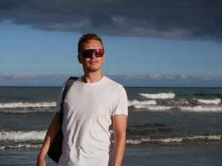 A young man with a backpack, wearing a white T-shirt and sunglasses standing on the seashore at the background of gloomy cloudy sky, with copy space. Tourism, traveling and resort recreation concept