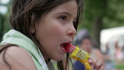 Girl enjoying a popsicle outdoors, wrapped in a towel. The scene reflects the joy of childhood and a refreshing summer treat in a lively park setting