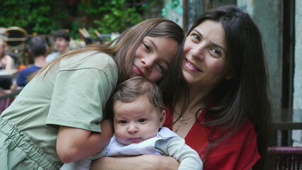 Obraz premium Mother and her two children sharing a warm embrace outdoors. Young girl hugs her mother and baby sibling, expressing love and closeness in a relaxed and joyful family moment
