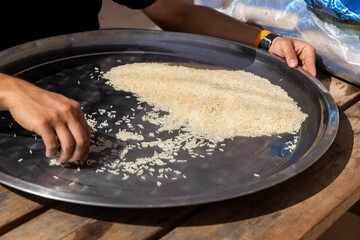 A man sorts rice on a big steel platter while seated at a wooden table, making pilaf.