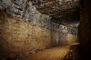 Obraz premium This image depicts a long underground stone tunnel featuring a wooden walkway on the right and rough stone walls illuminated by dim lights, showcasing medieval architecture.