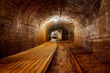 A dimly lit tunnel features a worn wooden walkway and rusty railroad tracks. The rough stone walls...