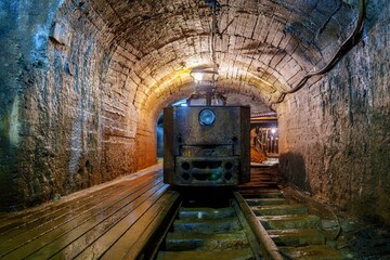 A rusty old mine locomotive sits idle in a dark mine tunnel with wooden tracks and stone walls. The...