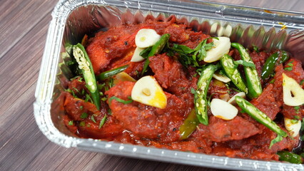 Overflowing with spicy, aromatic chicken dish, the fiery red sauce glistens with hints of garlic and green chilies sprinkled on top, surrounded by fresh herbs. It's served on a rustic wooden table.
