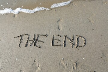 words THE END are large and engraved into the sand on the beach near the shore and a wave is coming