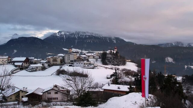 Picturesque winter scene of a snow-covered village in the Alps with traditional buildings, a church, and a mountain backdrop under cloudy skies.