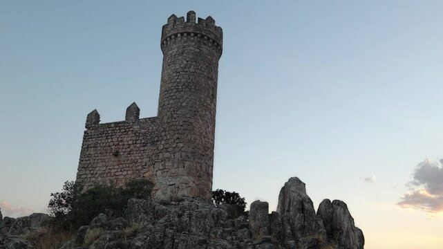 Captures an image of a rock castle with a tower on top, set on rock formations, with a sunset in the background and surrounded by trees.	