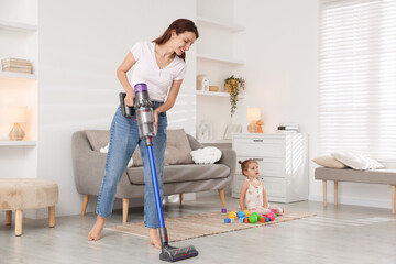 Smiling young woman cleaning floor with cordless vacuum cleaner while her daughter playing with...