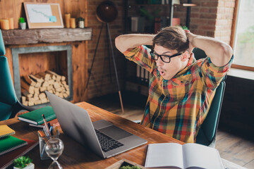 Photo of handsome young man wear checkered shirt sit hold arms behind head indoors office workplace workspace loft interior