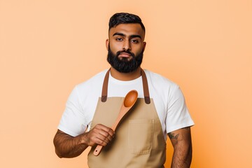 Serious Indian man in a beige apron holding a wooden spoon on a peach background, symbolizing confidence and culinary skills
