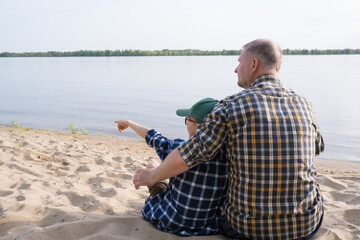 Father's day concept. Rear view of father and son sitting together on the river bank and looking at the water.