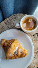 White ceramic cup with aromatic espresso and chocolate croissant on white plates and wooden table background with morning light. Coffee smile