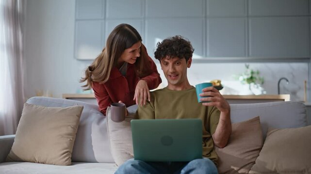 Home couple looking laptop at modern living room. Man sharing coffee with woman