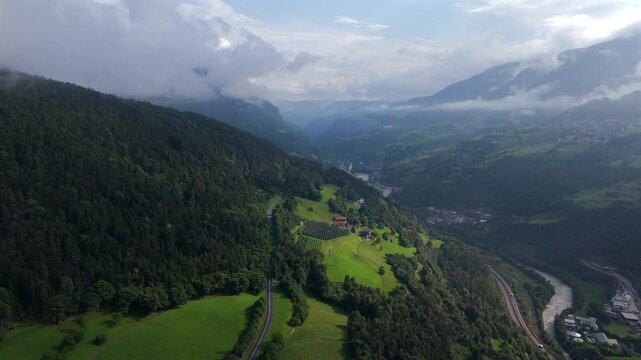 Aerial footage capturing a lush green valley with winding roads, dense forested hills, and a distant village nestled among the mountains in the Italian Alps.