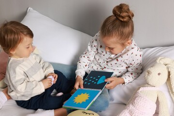 Sister and brother reading book together on bed indoors