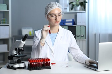 Laboratory testing. Doctor holding test tube with blood sample while working on laptop at table...