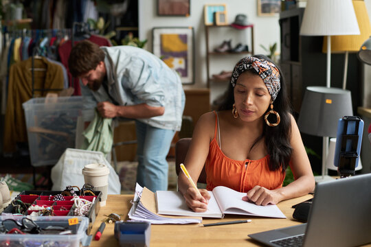 Young woman in stylish casualwear making notes in copybook while organizing work against male assistant with container