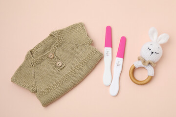 Pregnancy tests, rattle and baby clothes on beige background, top view