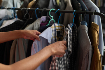 Hands of young unrecognizable female customer looking through secondhand casual attire on hangers...