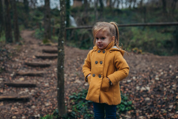A young child dressed in a yellow coat stands thoughtfully amidst a serene forest path,...