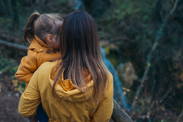 Fototapeta premium A parent and child, warmly dressed in yellow jackets, enjoy a peaceful moment together, standing beside a flowing forest stream, evoking feelings of warmth and love.