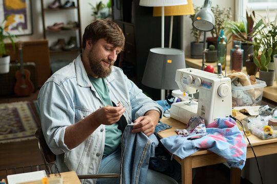 Smiling man sitting by table with sewing machine and stitching detail to denim jacket or jeans while upcycling secondhand apparel
