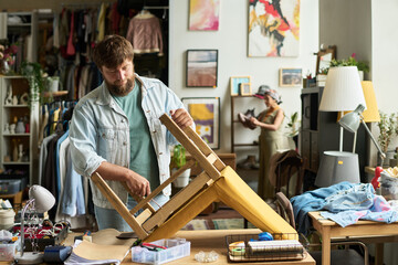 Bearded man with screwdriver fixing screws on bottom of secondhand wooden chair while working in...