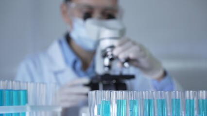 Laboratory tubes, filled with a blue liquid, are in focus, closeup view. Female scientist researcher wearing white gloves and protective mask is using a microscope at the background