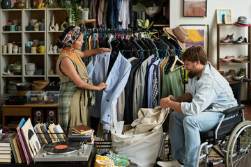 Young male buyer in wheelchair choosing secondhand attire from sack against woman standing by rack with new collection