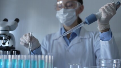Scientist woman, wearing a lab coat, mask, blue gloves and safety glasses, is using a micropipette...